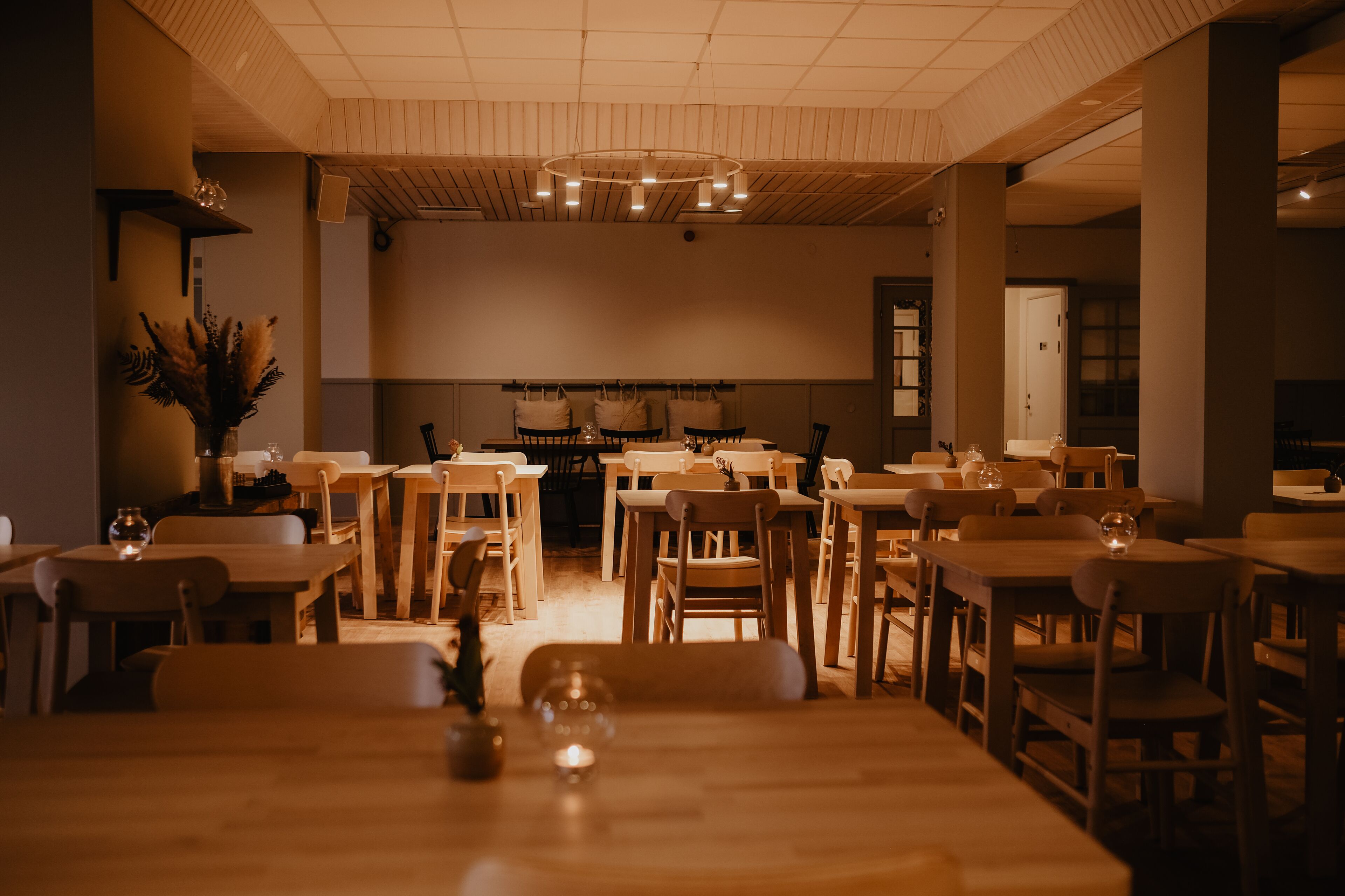 A spacious dining area with multiple wooden tables and chairs, a central chandelier, and a decorative plant on the left.