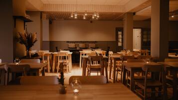 A spacious dining area with multiple wooden tables and chairs, a central chandelier, and a decorative plant on the left.