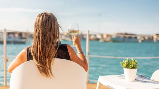 Une personne assise à une table au bord de l’eau, tenant un verre de vin blanc.