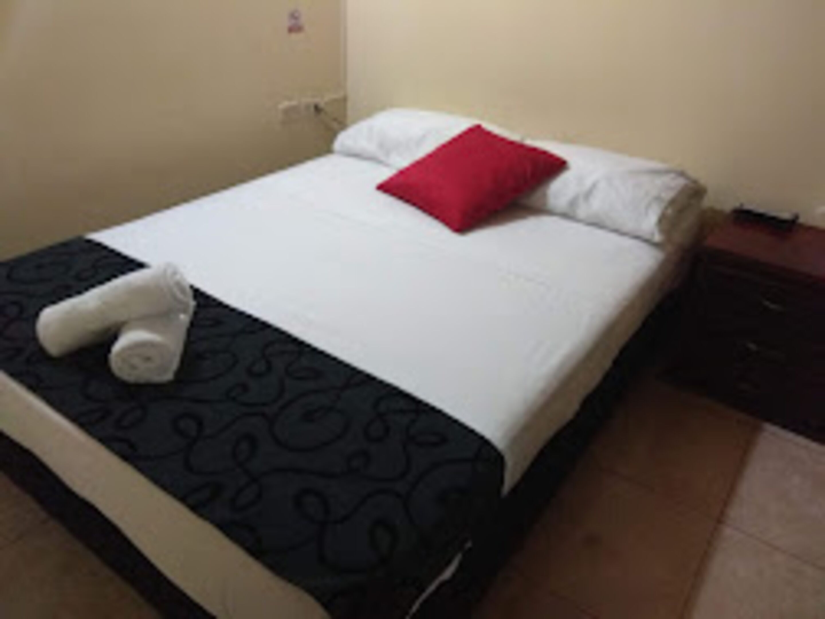 Een net opgemaakt bed met een zwart-witte sprei, een rood kussen en opgerolde handdoeken aan de zijkant.