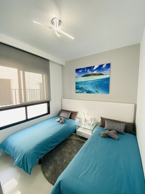 A modern bedroom with two beds, a nightstand, a ceiling fan, and a beach-themed painting.