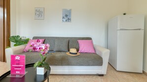 Una sala de estar con un sofá gris, cojines rosas, una pequeña mesa con una orquídea en maceta, un cuadro enmarcado y un refrigerador blanco.