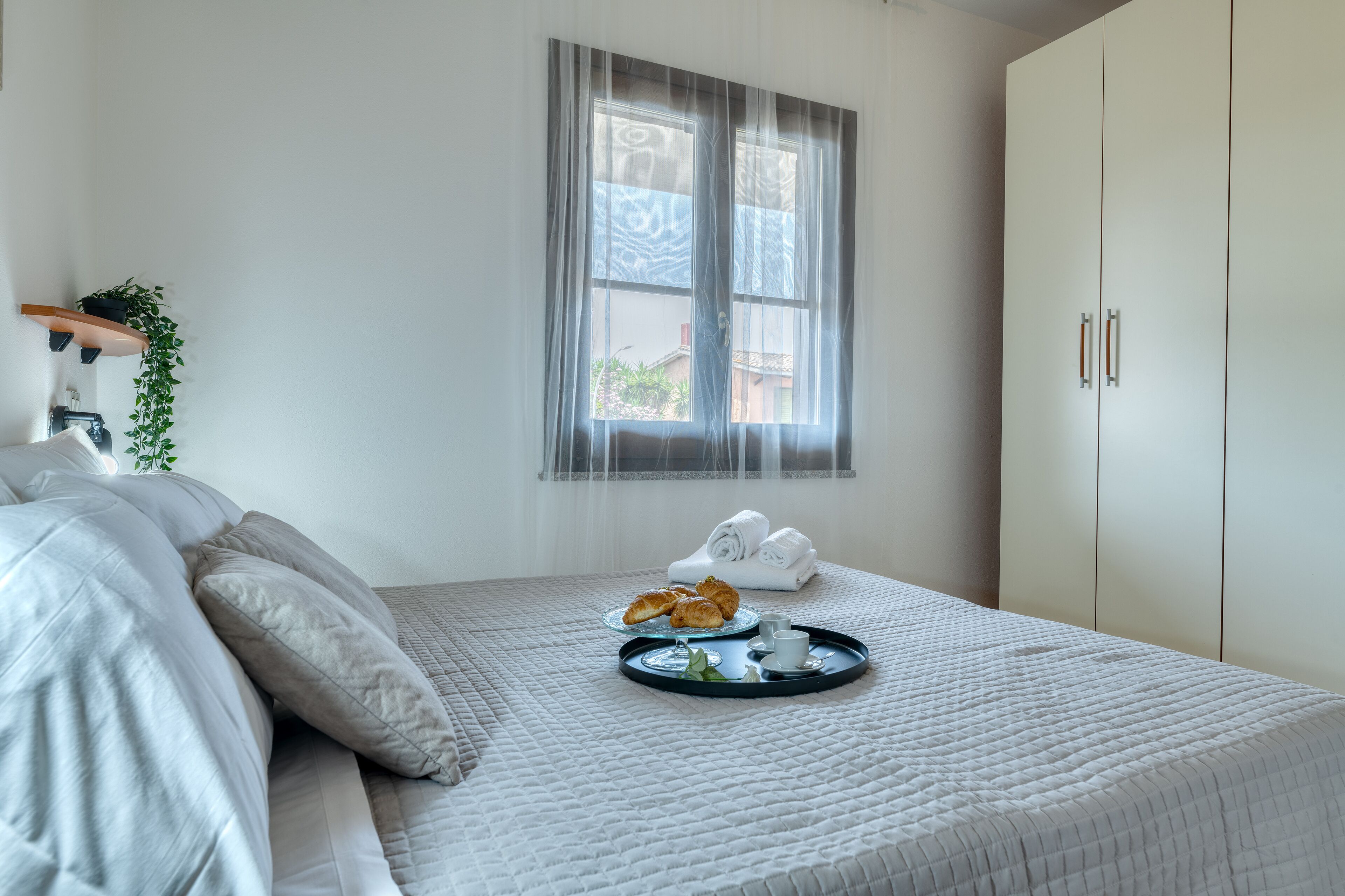 Una cama bien hecha con almohadas, una bandeja con croissants y una taza, y una ventana con cortinas traslúcidas.