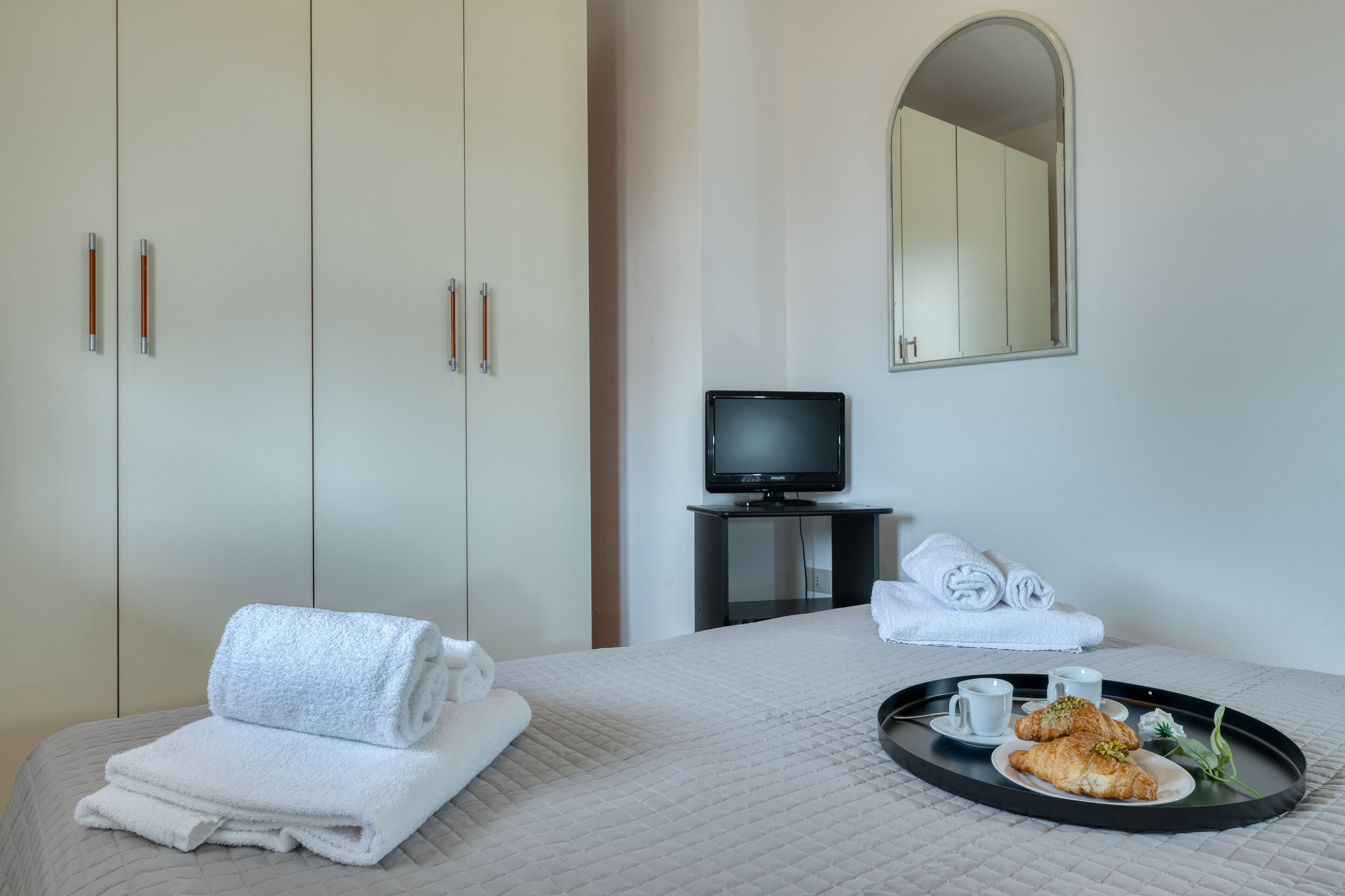 A hotel room with a bed, a television on a stand, a tray with croissants and a cup, and a mirror on the wall.