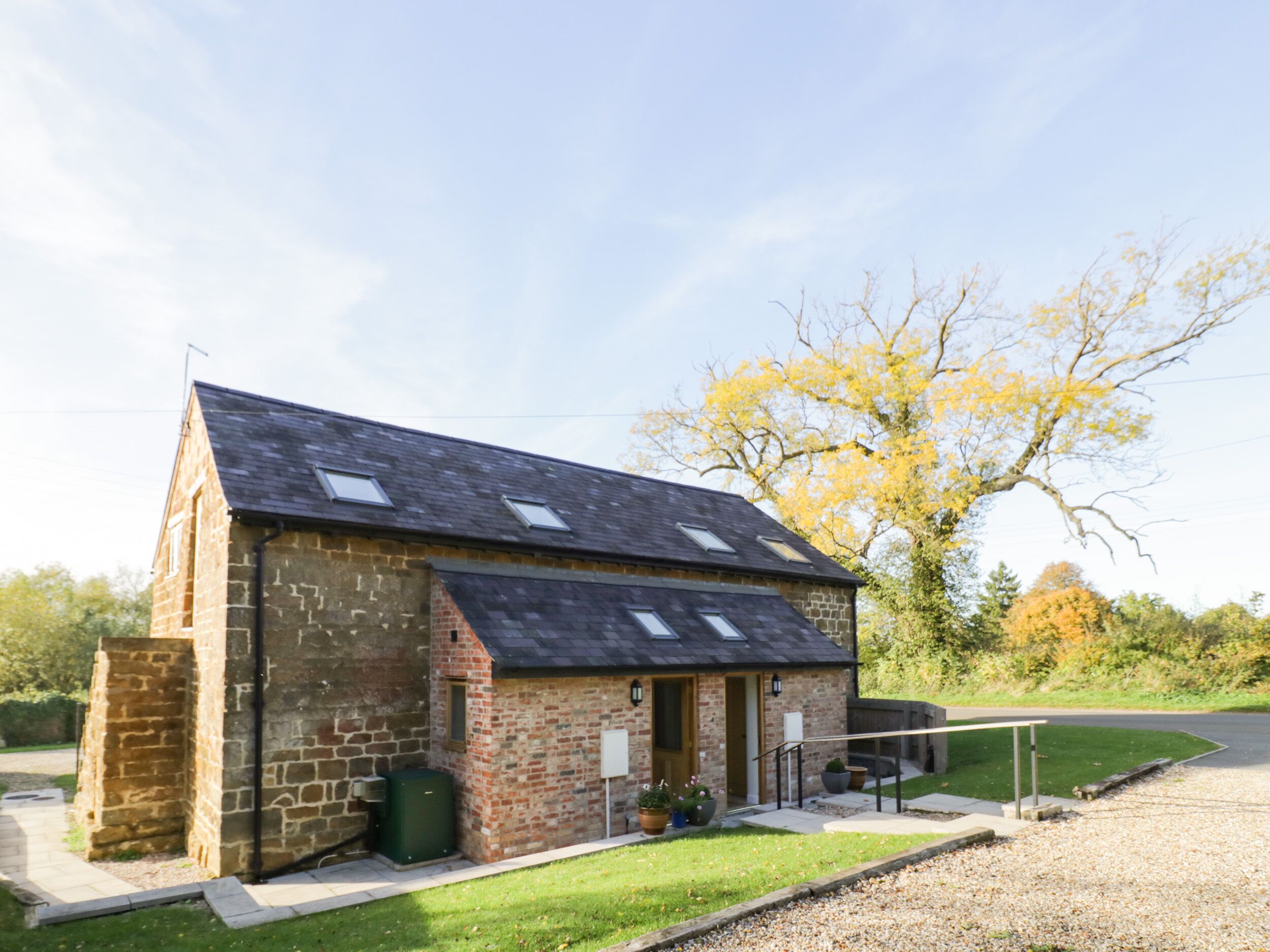 A small stone building with a sloped roof and skylights, a paved entrance, and a grassy area with a tree.