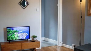 A living room with a TV on a wooden stand, a floor lamp, and a door leading to another room. - George's City Centre Apartment - Salisbury (Salisbury)