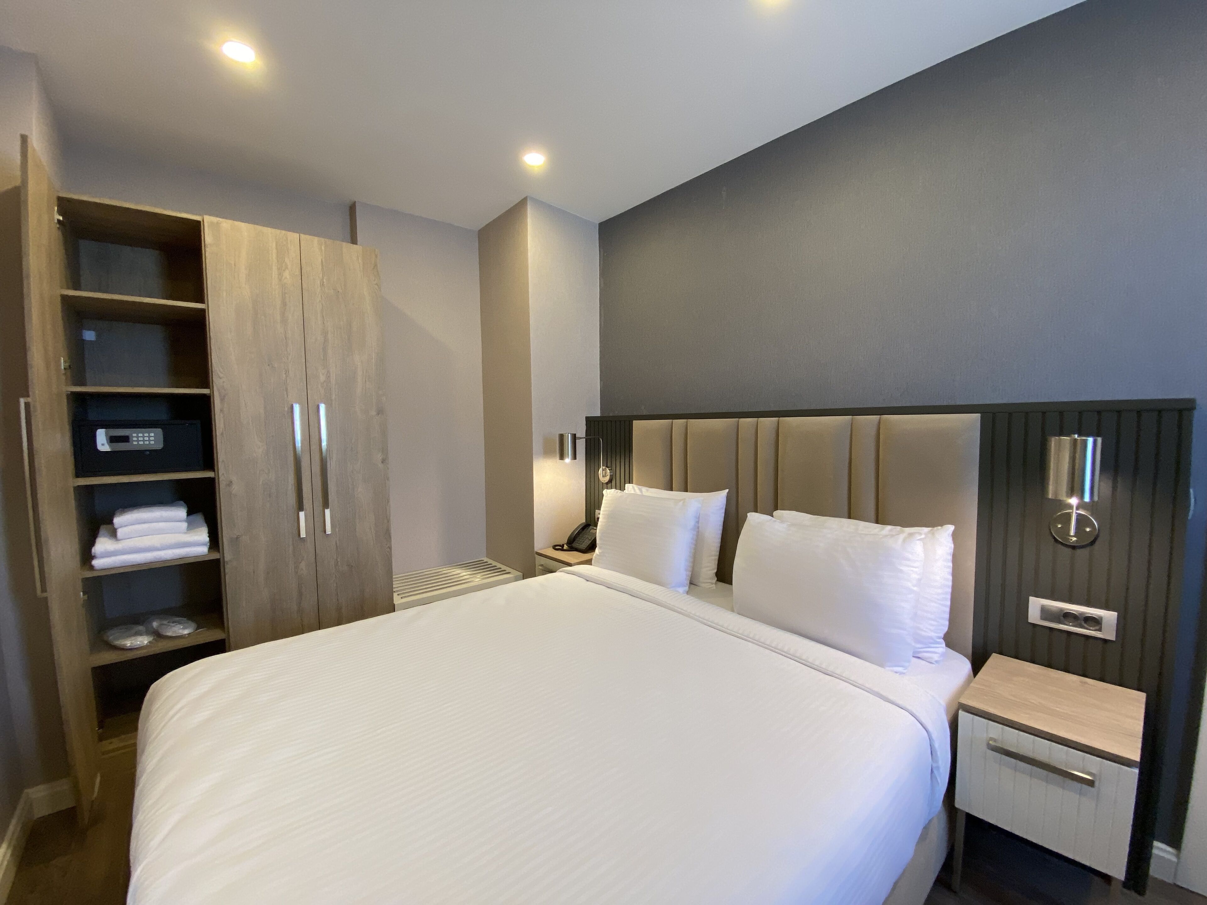 A modern hotel room with a large bed, a wooden wardrobe, a bedside table, and a nightstand.