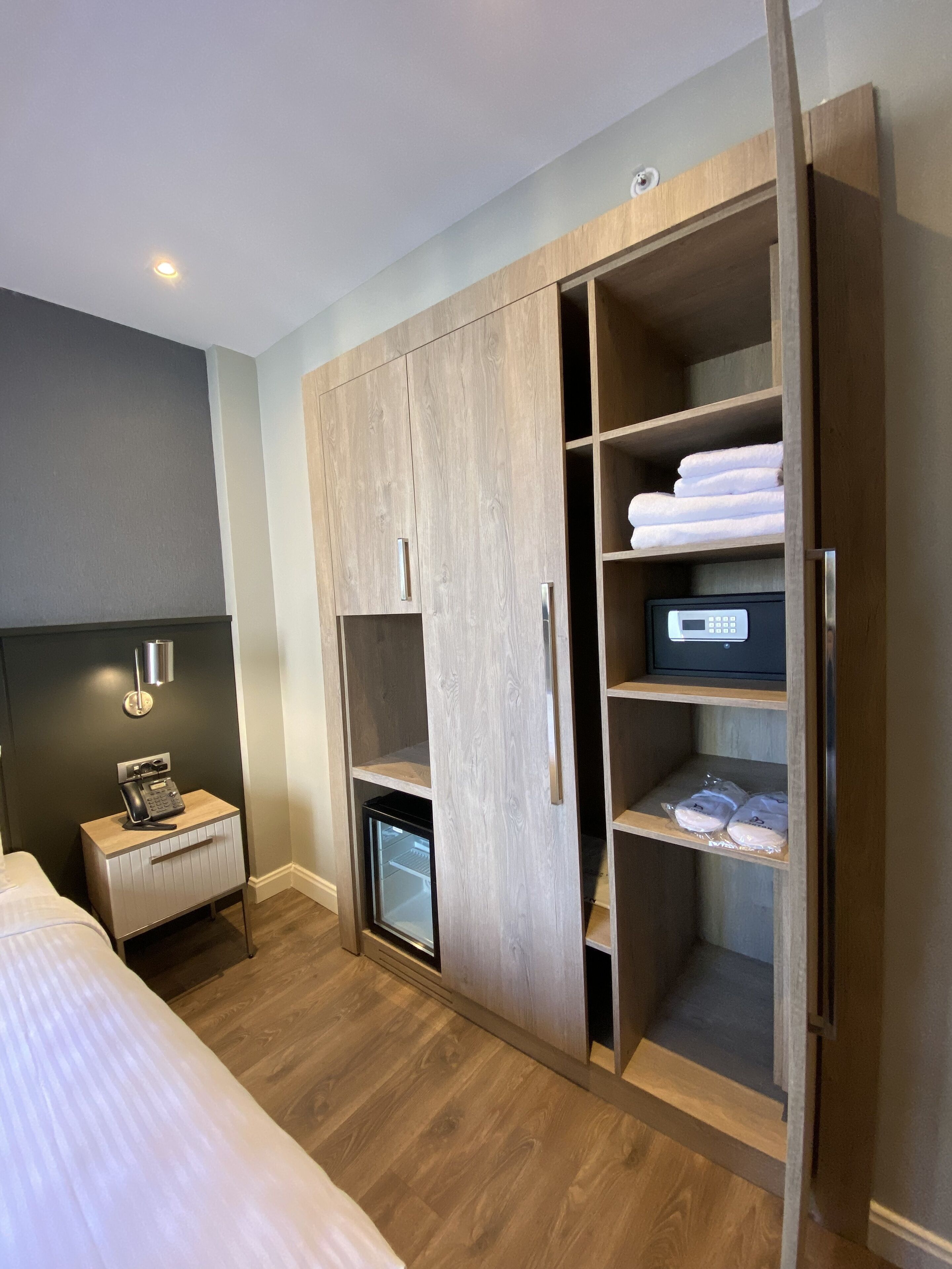 A hotel room with a wooden wardrobe, a bedside table with a phone, and a small refrigerator.