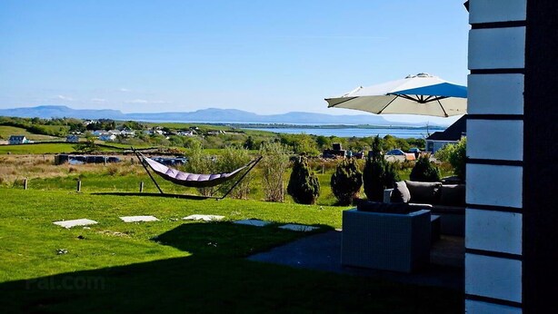 A view of a grassy area with a hammock and outdoor furniture, a white building with a striped facade, and a distant landscape with mountains and a body of water.