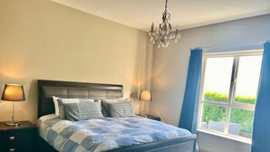 A bedroom with a bed, two bedside tables, a bench, a chandelier, and a window with curtains.