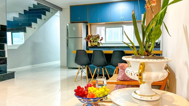 Une cuisine moderne avec des meubles bleus, un réfrigérateur, un coin repas avec des chaises noires et une table agrémentée d’un panier de fruits et d’un vase de fleurs.