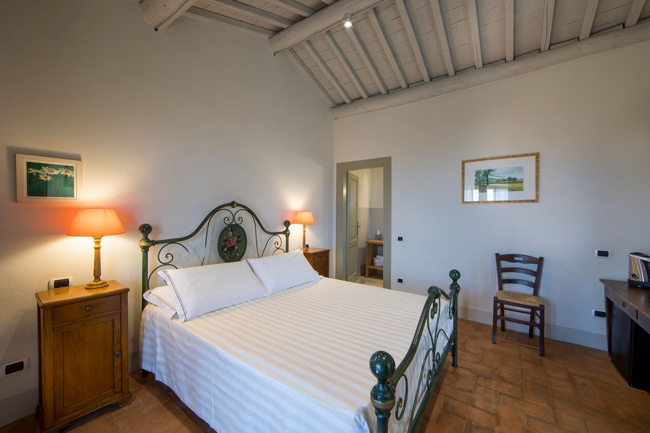 Quarto com cama, duas luminárias de cabeceira, mesa de cabeceira de madeira, uma cadeira e uma escrivaninha.