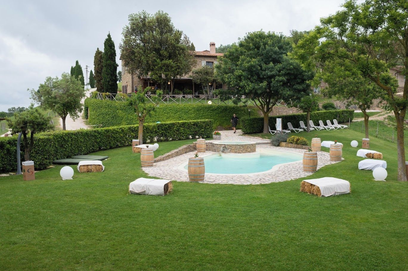 Zona de piscina con solárium de piedra, rodeada de césped y árboles, con sillones de descanso y fardos de heno.