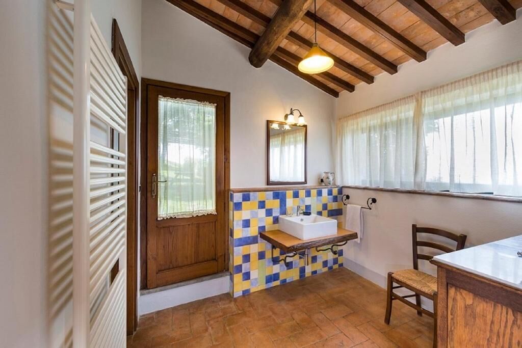 Un baño con puerta de madera, salpicadero de azulejos, lavamanos, espejo y ventana con cortinas.