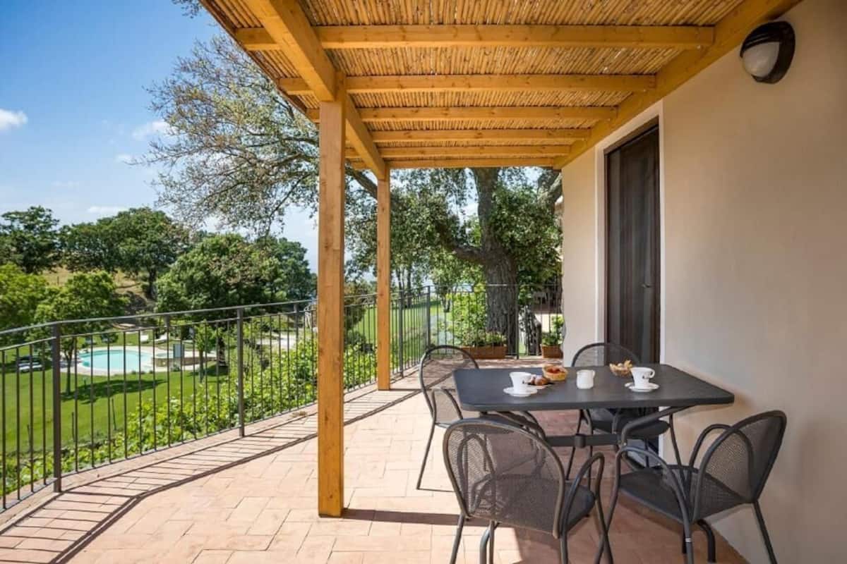 Een overdekt terras met een tafel en stoelen, een zwembad in de verte en een omheinde tuin.
