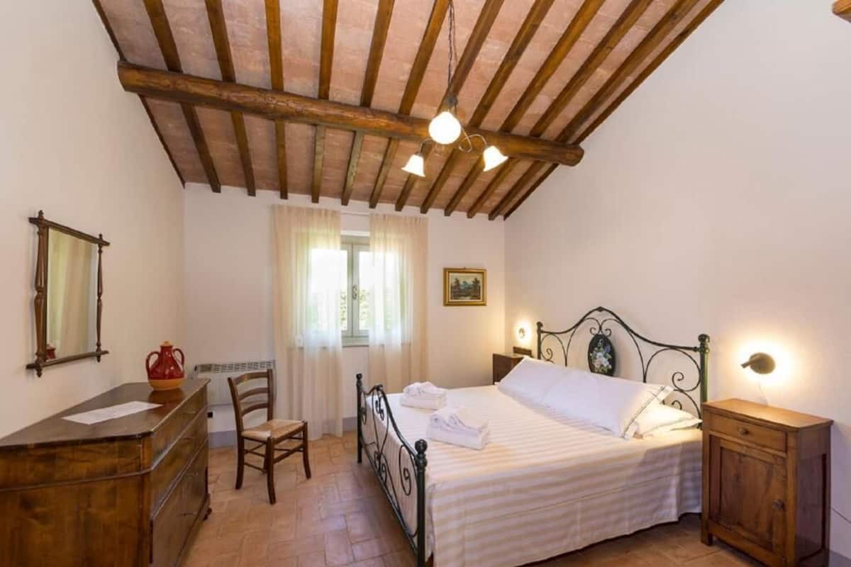 Een slaapkamer met een houten plafond, een bed met wit beddengoed, een bureau met een spiegel en een stoel.