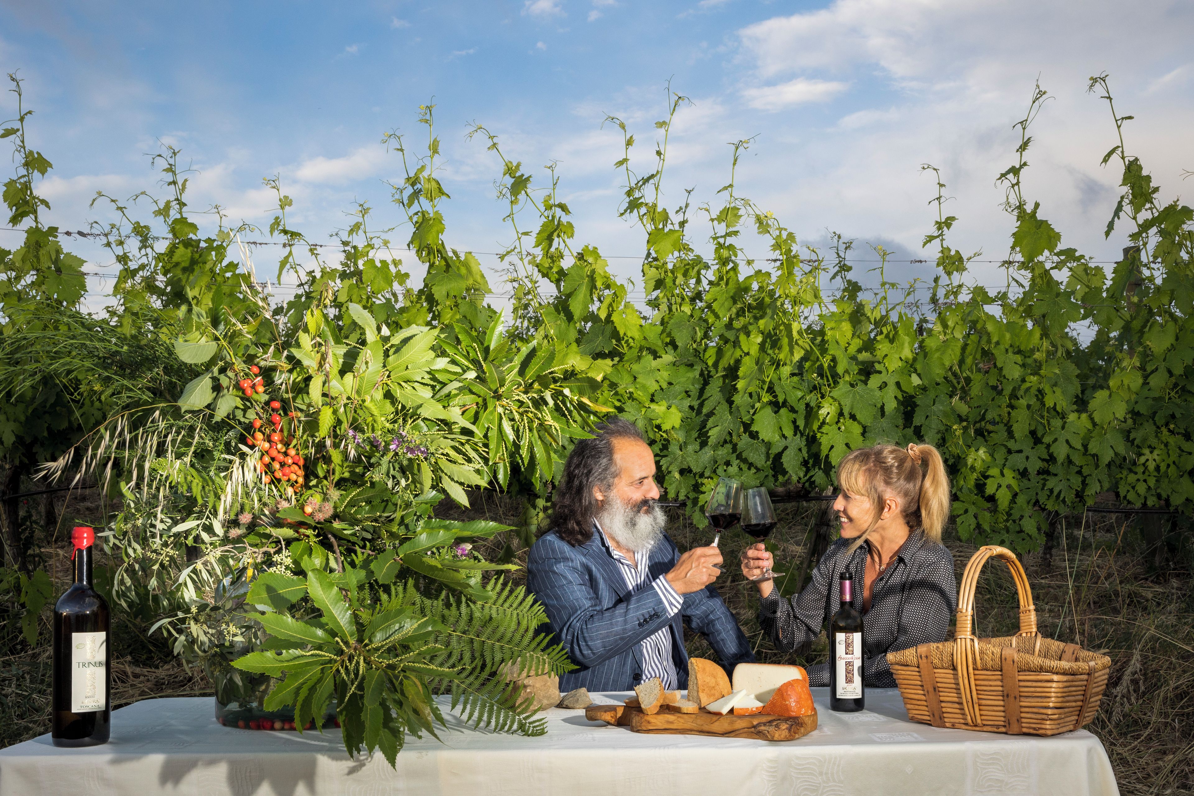 Two people seated at a table with a bottle of wine, bread, and a basket, surrounded by a vineyard.