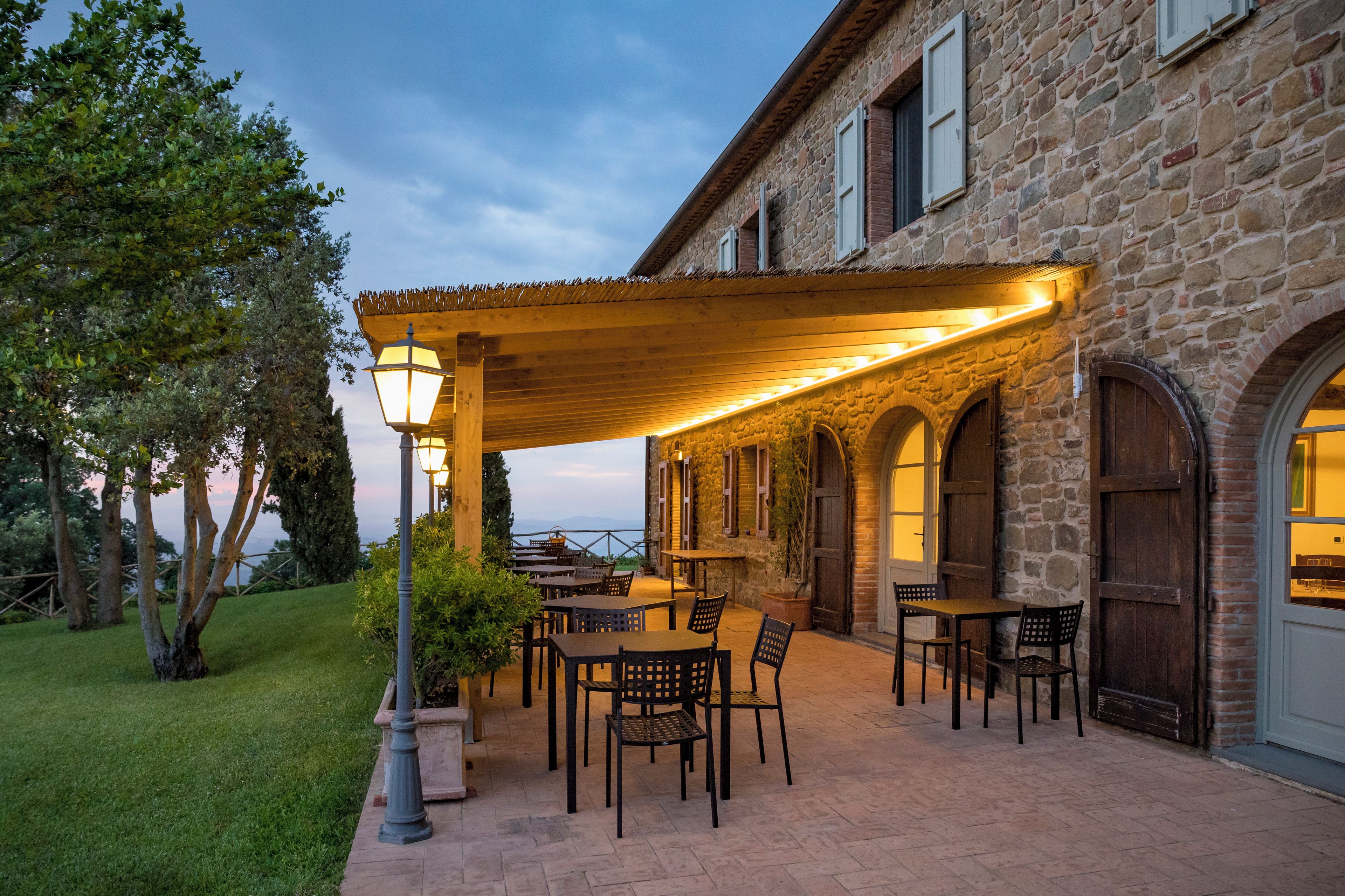 An outdoor patio with tables and chairs, a stone building with arched windows, and a view of the sea.