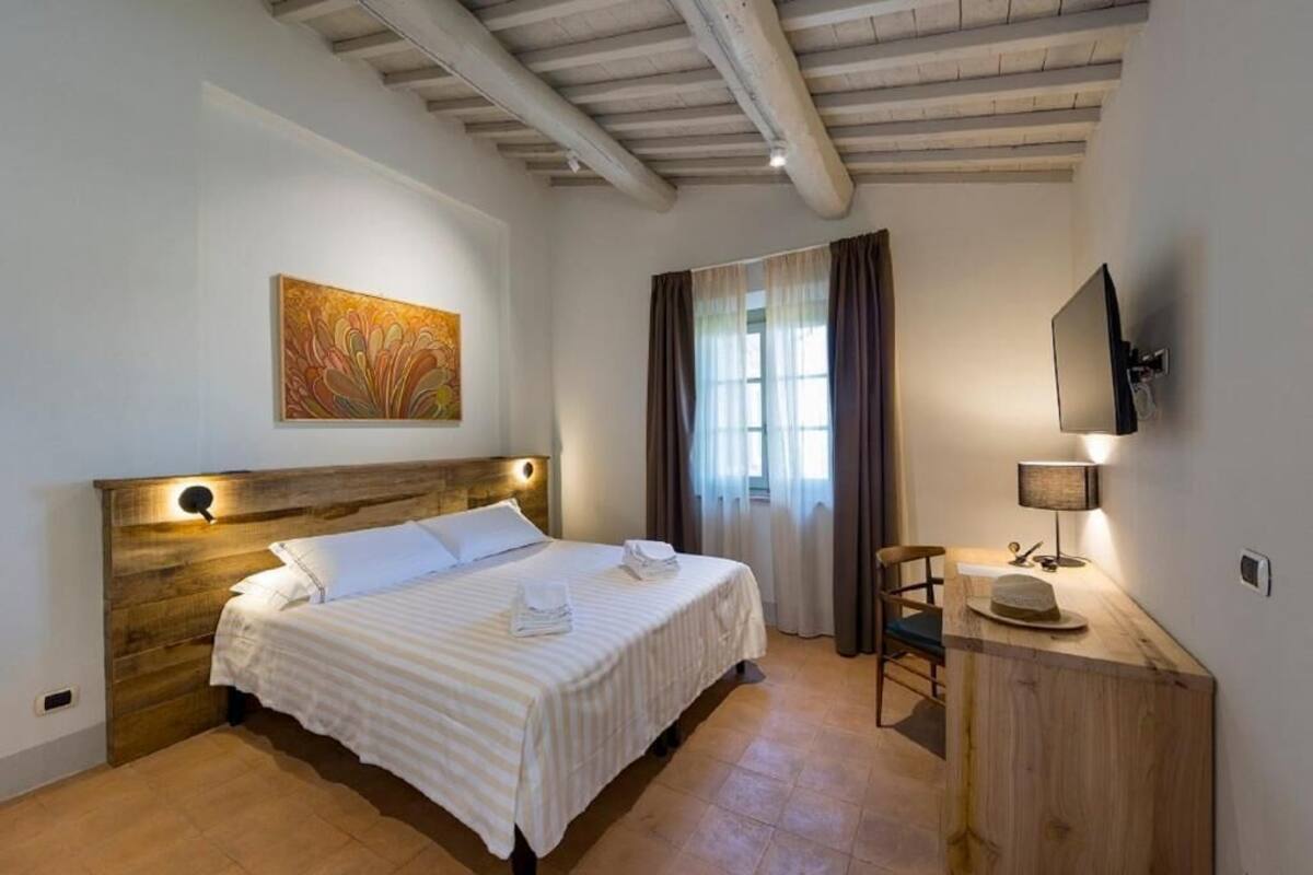 Een hotelkamer met een houten hoofdeinde, een bed met witte lakens, een schilderij aan de muur, een aan de muur bevestigde televisie en een bureau met een lamp en een hoed.