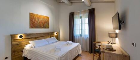 A hotel room with a wooden headboard, a bed with white linens, a painting on the wall, a TV mounted on the wall, and a desk with a lamp and a hat.