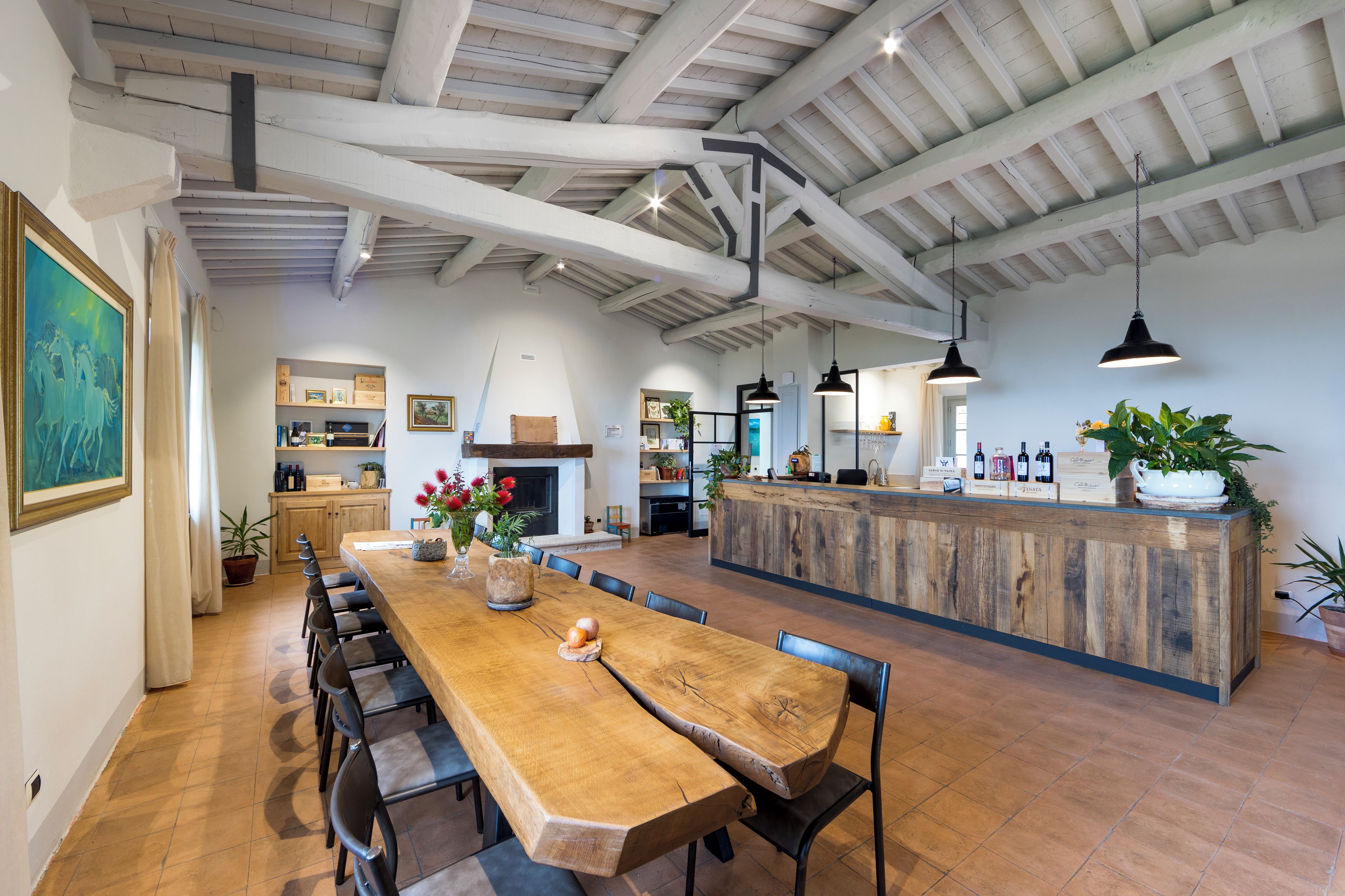 A spacious dining area with a long wooden table and chairs, a kitchen in the background, and a bar area with bottles and plants.