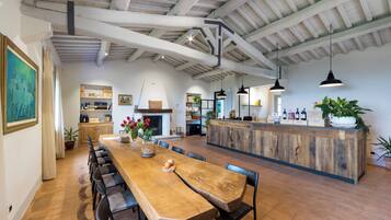 A spacious dining area with a long wooden table and chairs, a kitchen in the background, and a bar area with bottles and plants.
