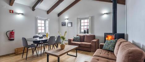 A spacious living area with a high ceiling, wooden beams, a fireplace, a dining table with chairs, and a sofa.