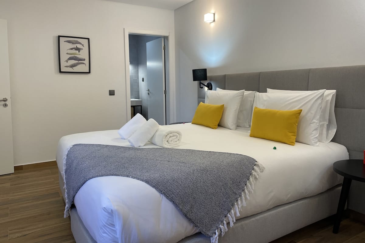 Una cama bien hecha con almohadas blancas y amarillas, cabecera gris y una manta gris.