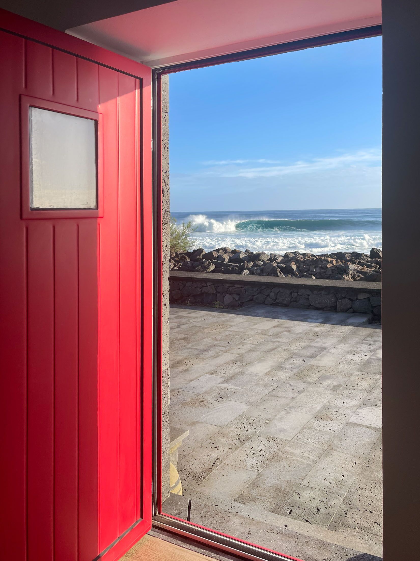 Una puerta roja que da a un balcón con vistas al océano y un muro de piedra.