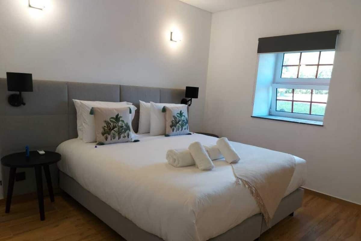 Una cama bien hecha con sábanas blancas y un cojín decorativo, una mesita de noche con una lámpara, y una ventana con vistas a la vegetación.