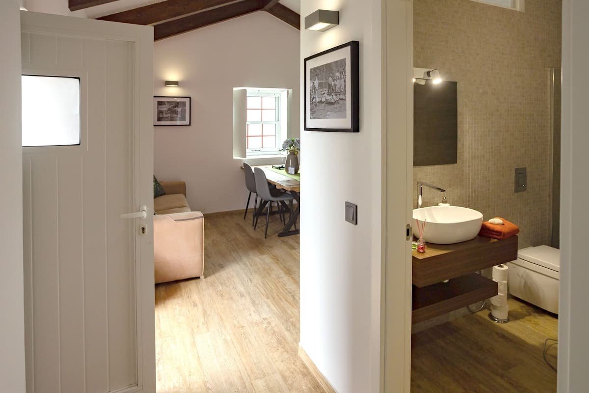 Una habitación con piso de madera, una puerta blanca, un área de comedor con sillas y una mesa, y un baño con lavamanos e inodoro.