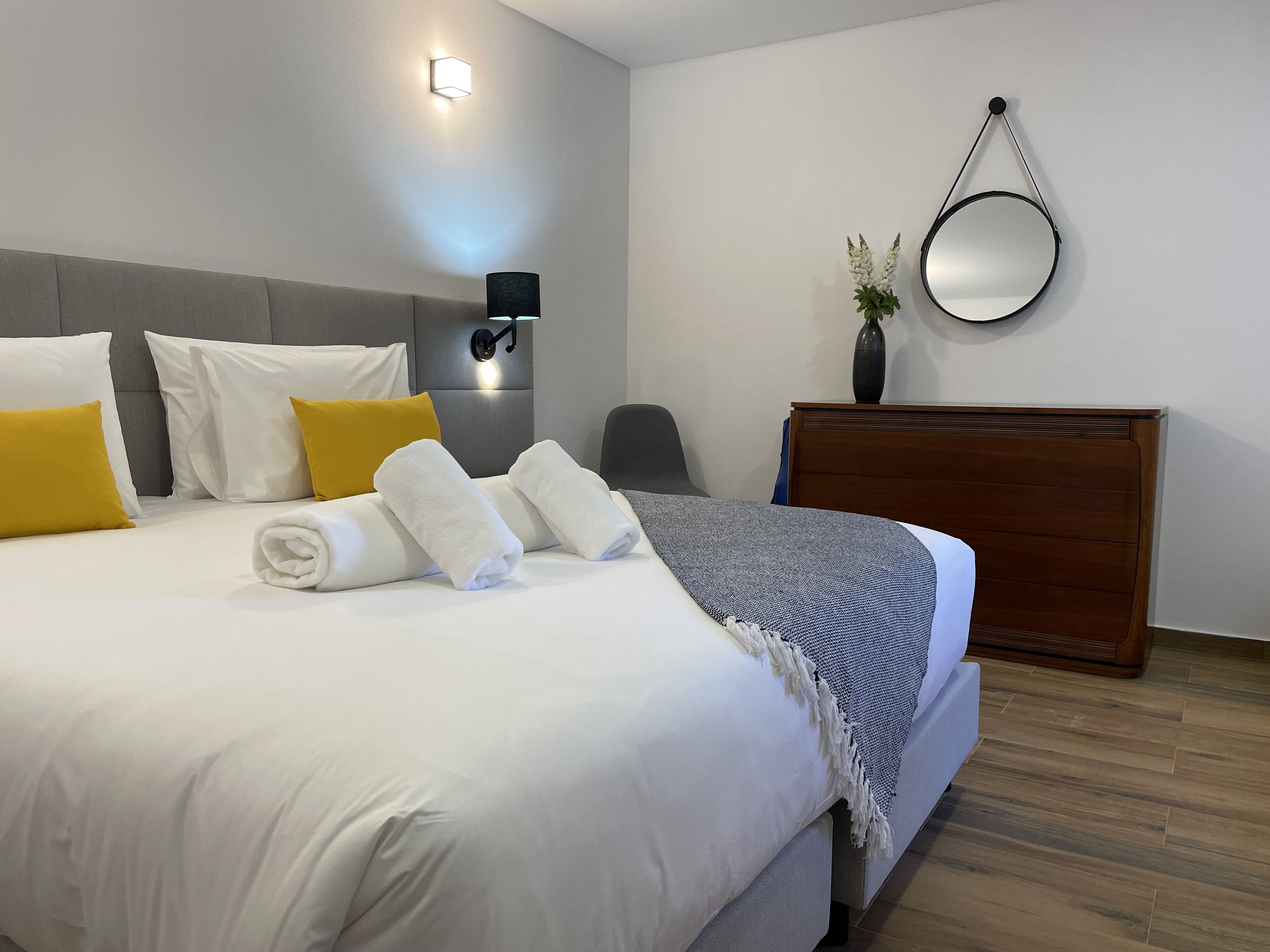 A neatly made bed with white and yellow pillows, a grey headboard, and a bedside table with a lamp.