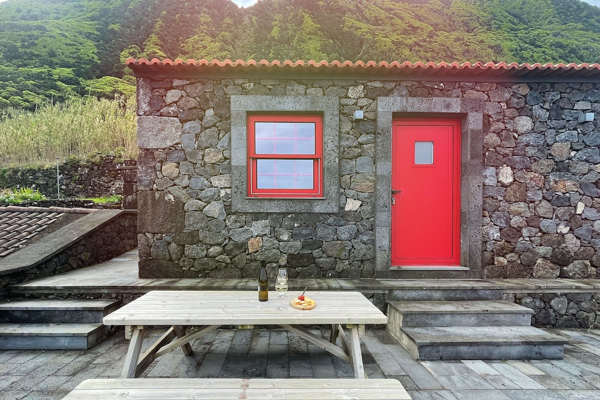 Un edificio de piedra con puerta y ventana rojas, una mesa de picnic y una botella de vino.