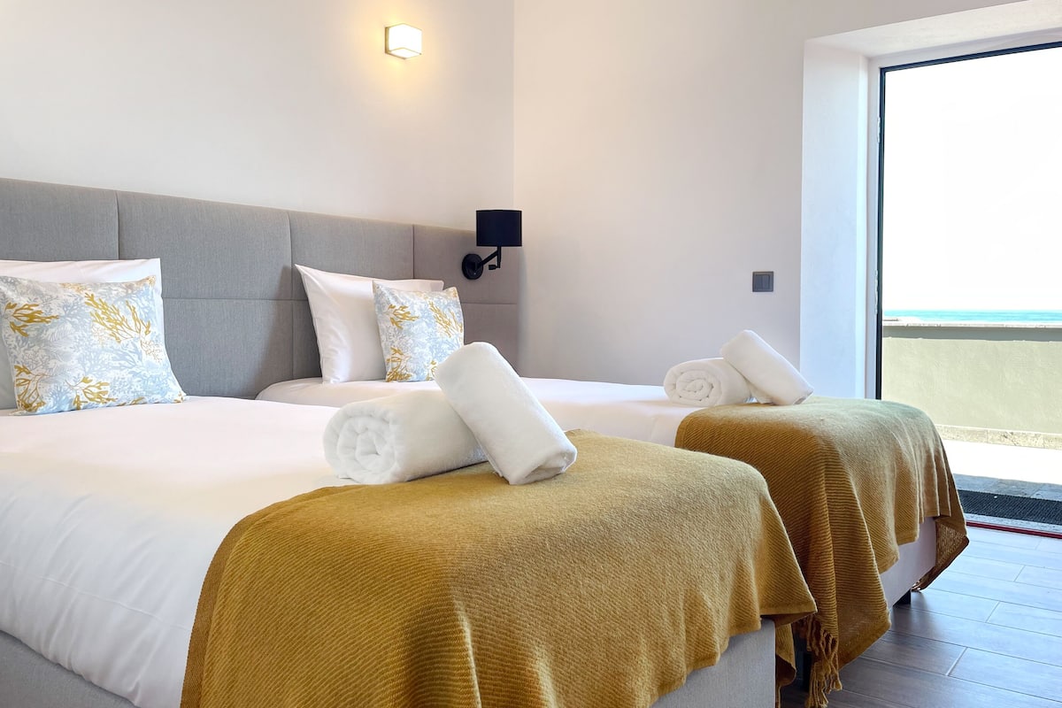 Habitación de hotel con dos camas, cada una con una manta blanca y un plaid color mostaza. Hay una lámpara de pared y una ventana con vista a la playa.