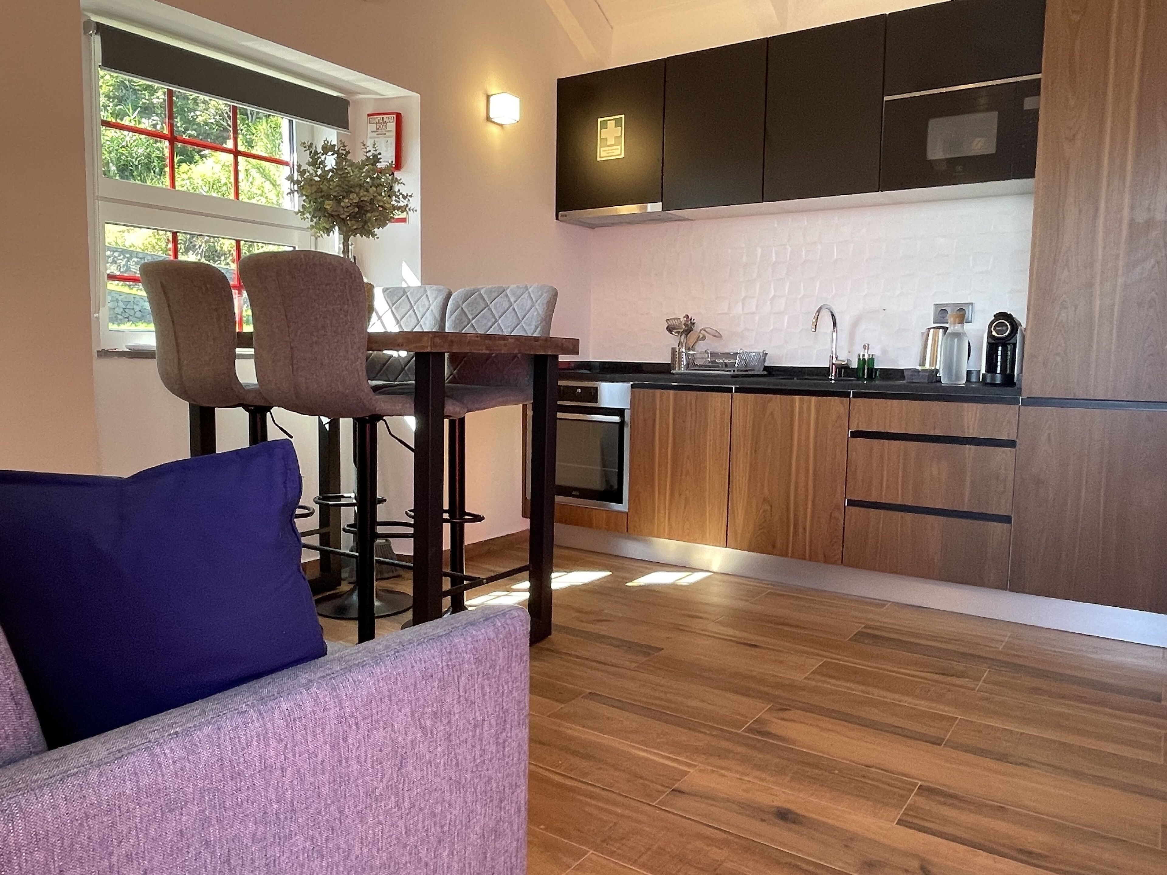 A modern kitchen with wooden cabinets, a built-in oven, and a sink. There is a dining area with a table and chairs, and a window with a view of greenery outside.