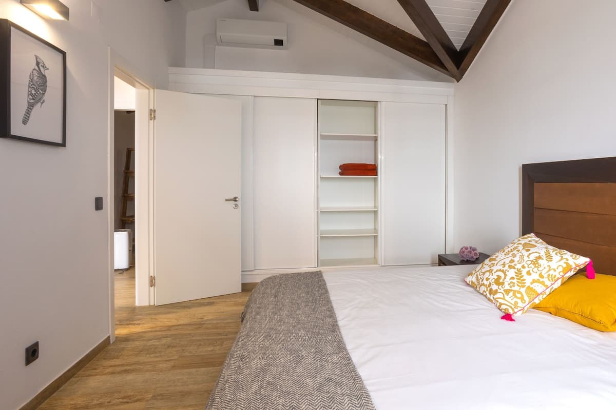 Un dormitorio con techo de madera, una cama blanca con almohadones estampados, un armario blanco y suelo de madera.