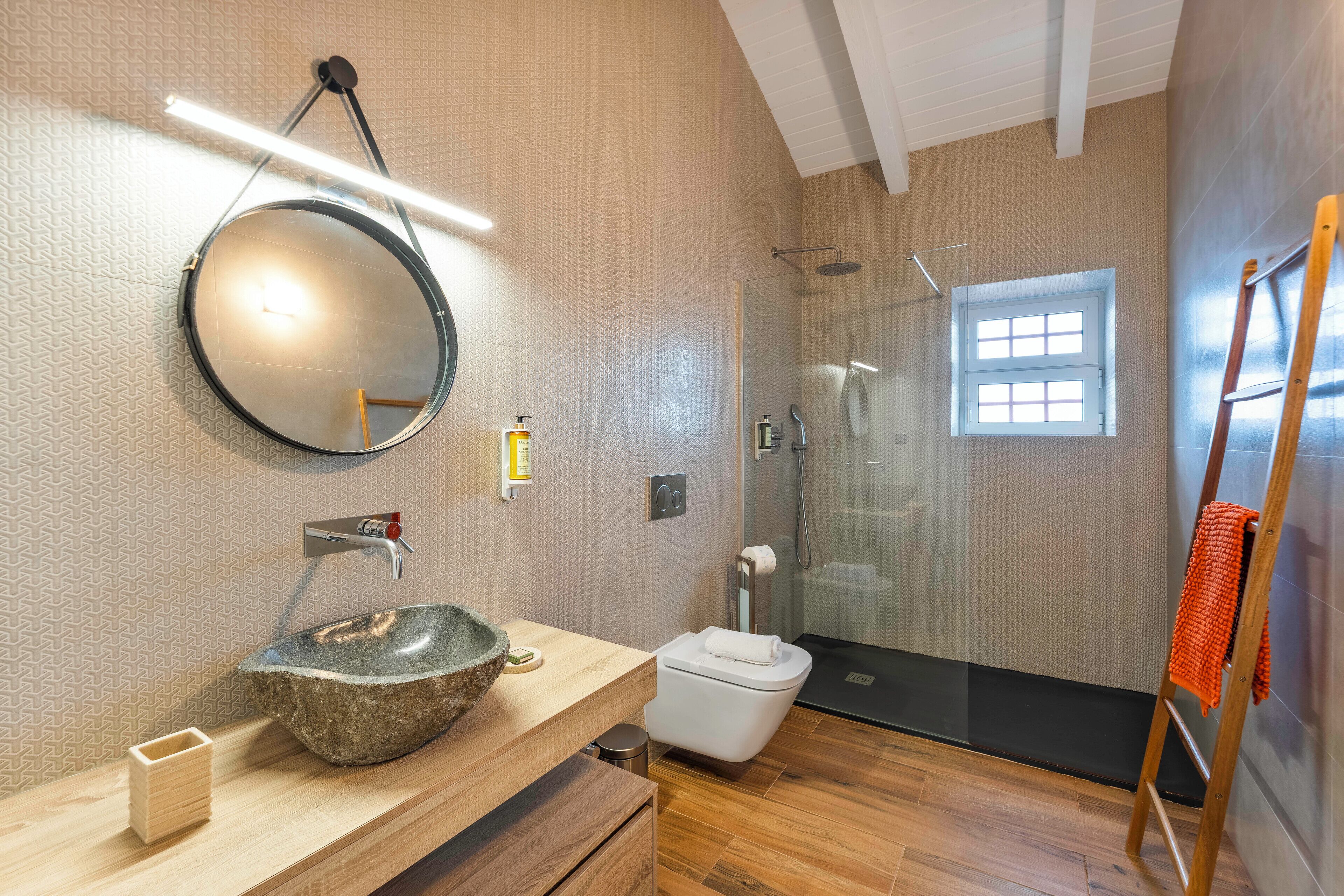 A modern bathroom with a stone sink, a round mirror, a glass shower enclosure, and a wooden ladder.
