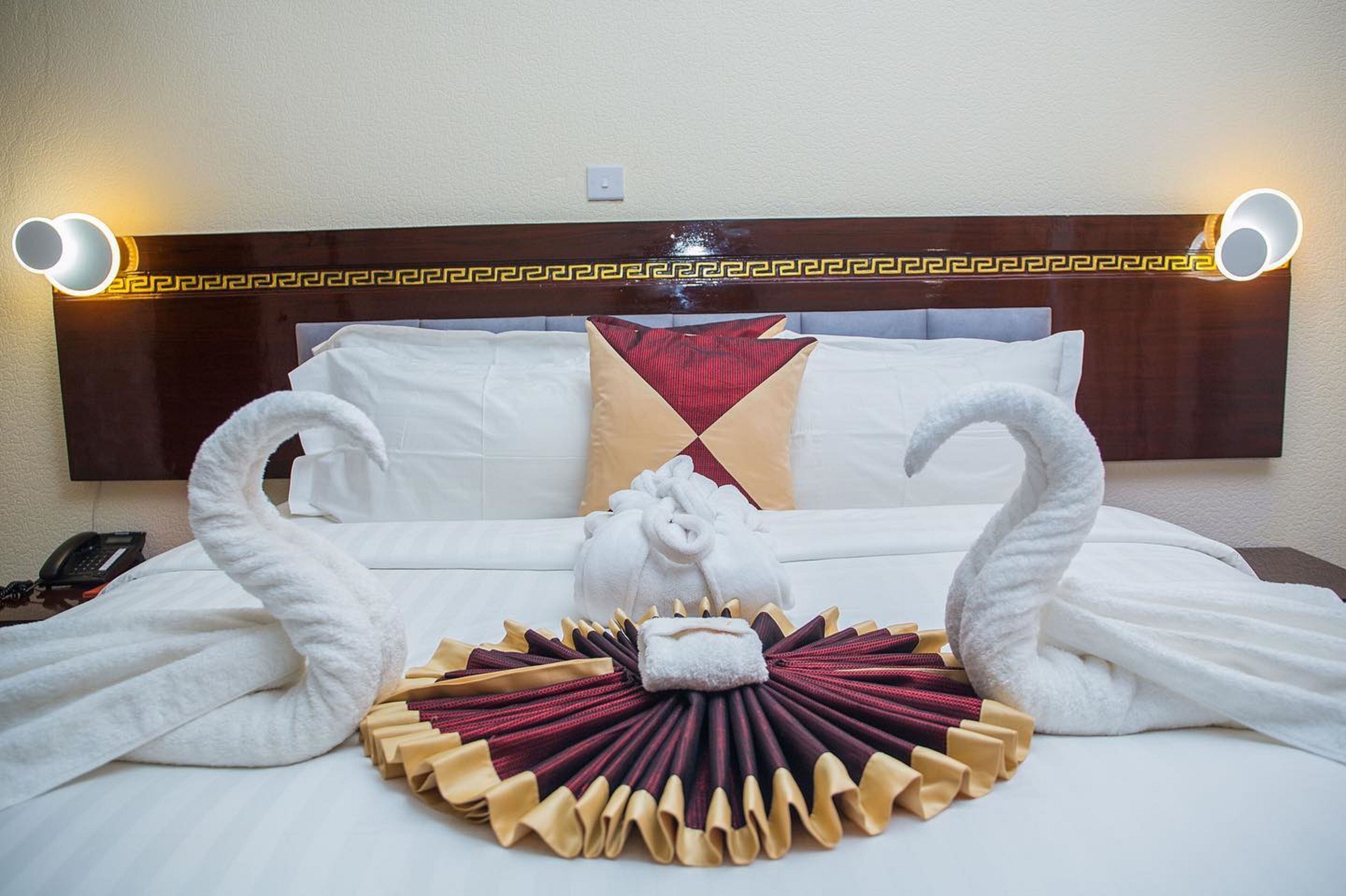 A hotel room with a bed featuring a decorative towel arrangement in the shape of swans, a headboard with a geometric pattern, and wall-mounted lights.