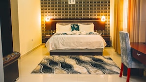 A hotel room with a bed, a desk, a chair, a patterned rug, and a wall with a geometric design and framed pictures. - Lion Luxury Suites (Enugu)