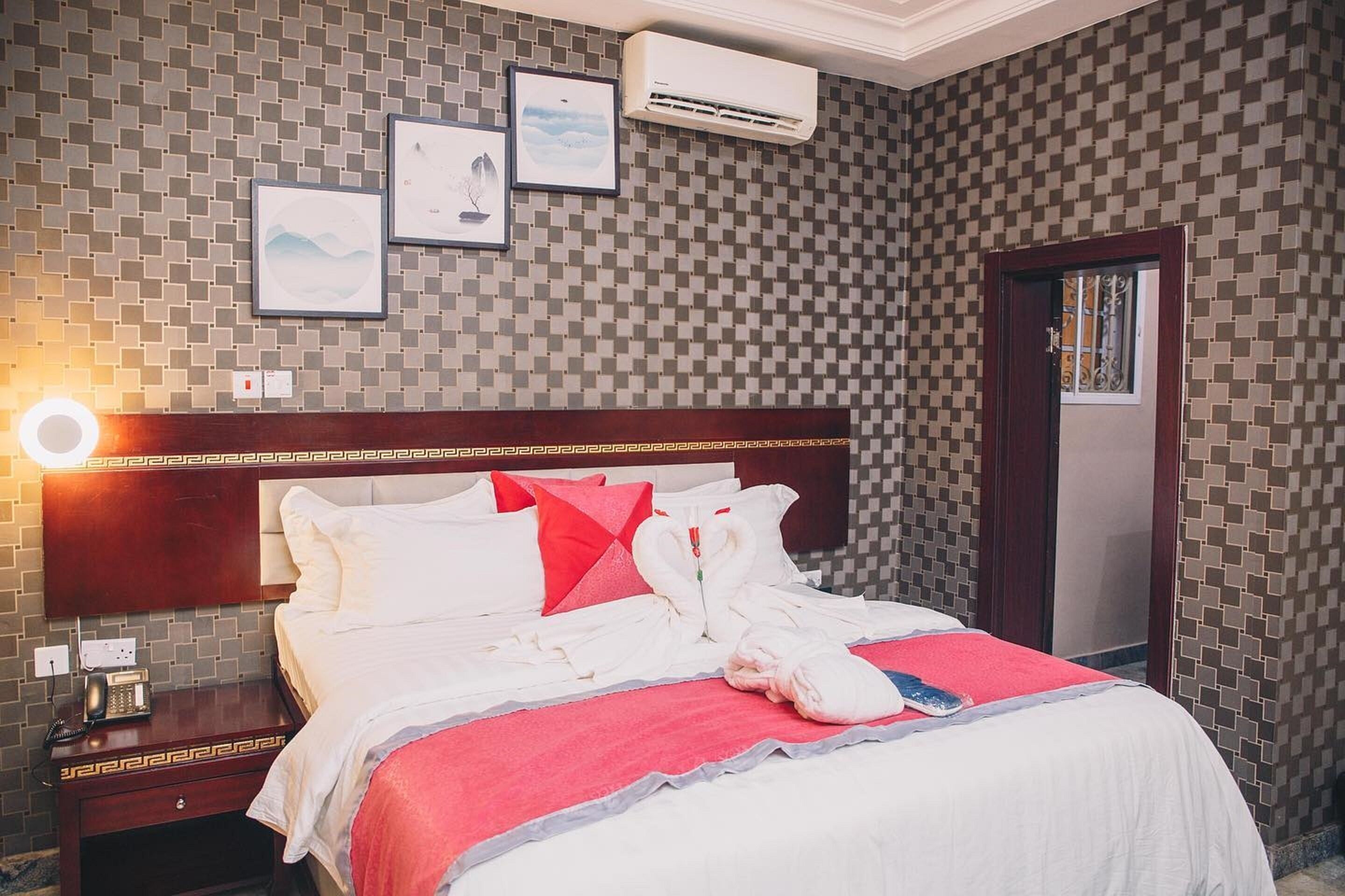 A hotel room with a checkered wall pattern, a wooden headboard, a bed with white bedding and red accents, a bedside table with a phone, and framed pictures on the wall.