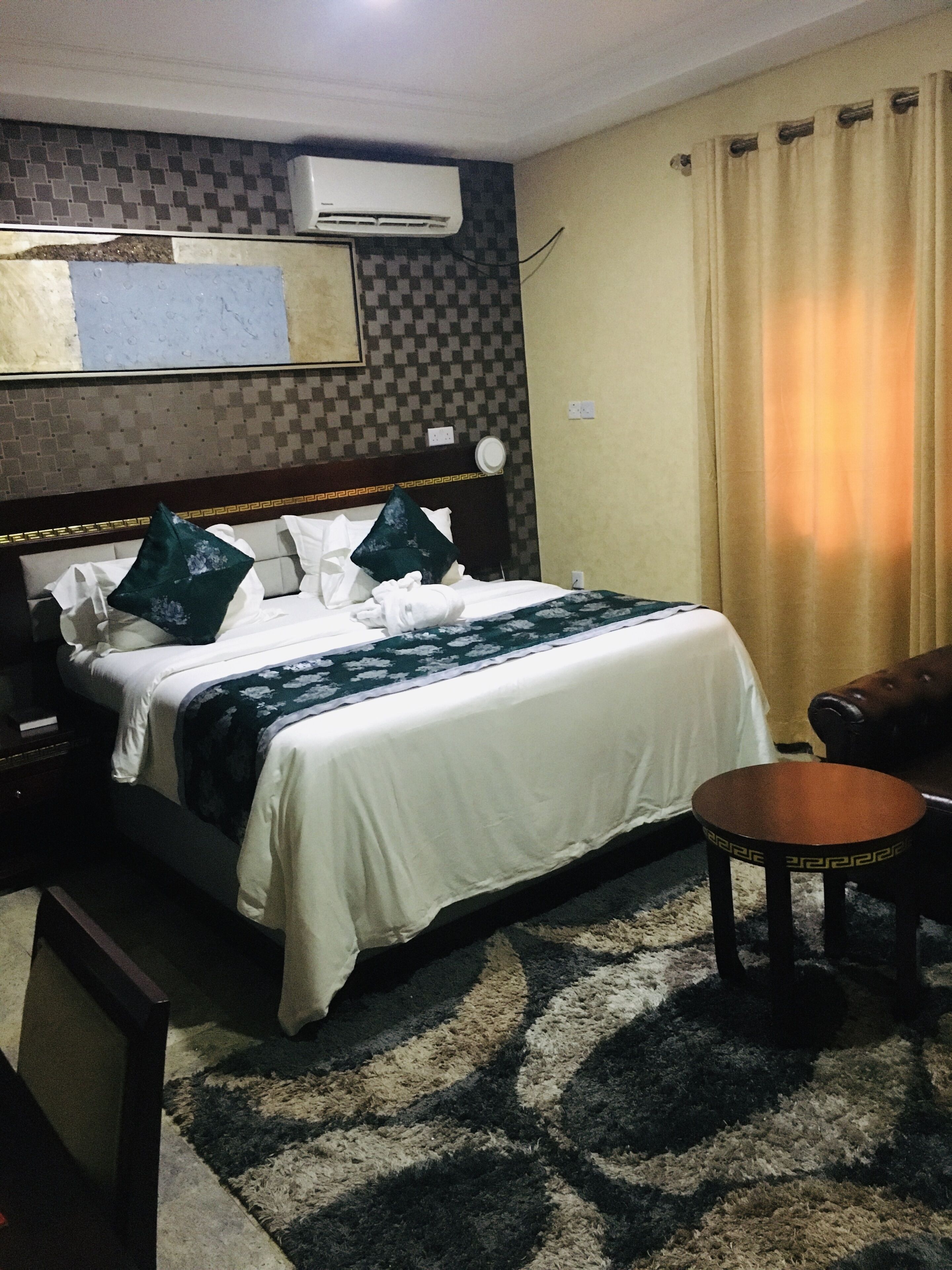 Quarto de hotel com cama, mesa redonda pequena, sofá, unidade de ar condicionado e tapete estampado.