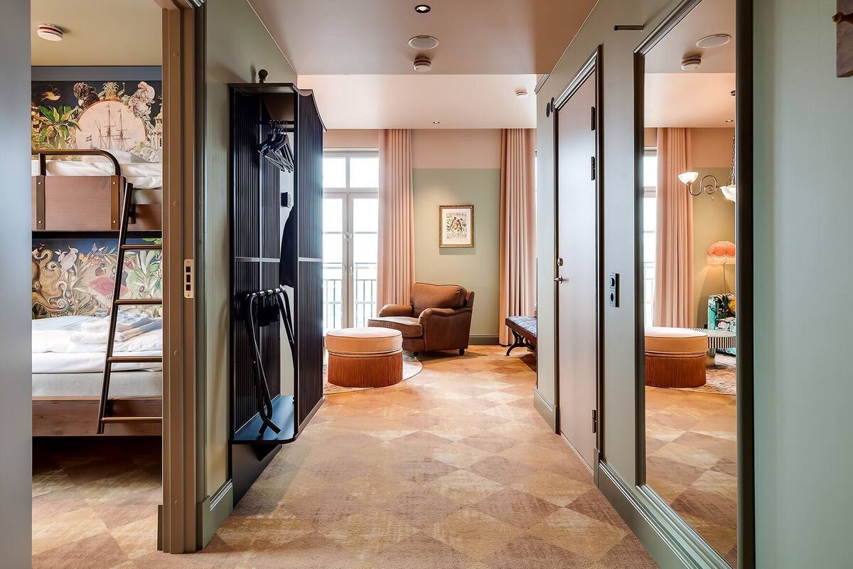 Une chambre d’hôtel comprenant un lit, un coin salon avec un fauteuil et un pouf, une armoire et une salle de bain visible à travers une porte ouverte.