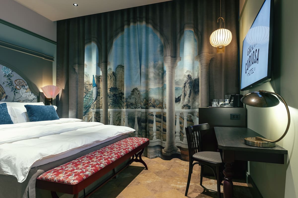 Une chambre d’hôtel avec un grand lit, un bureau, une chaise, un banc et une vue sur un espace extérieur agréable, visible par la fenêtre.