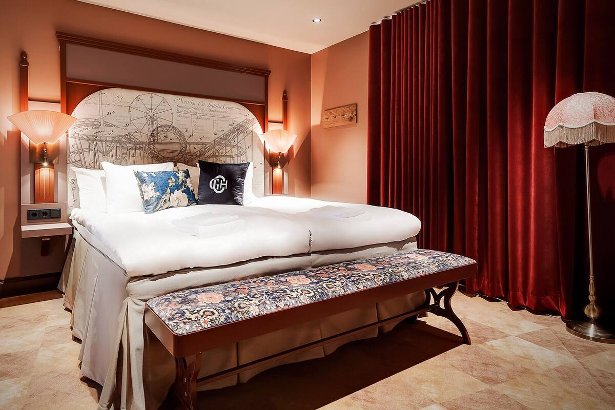 Une chambre d’hôtel avec un grand lit, une banquette, une table de chevet et une lampe fixée au mur.