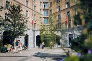 Entrada de hotel com uma placa indicando "CURIOSA HOTEL" e uma grande árvore na frente.
