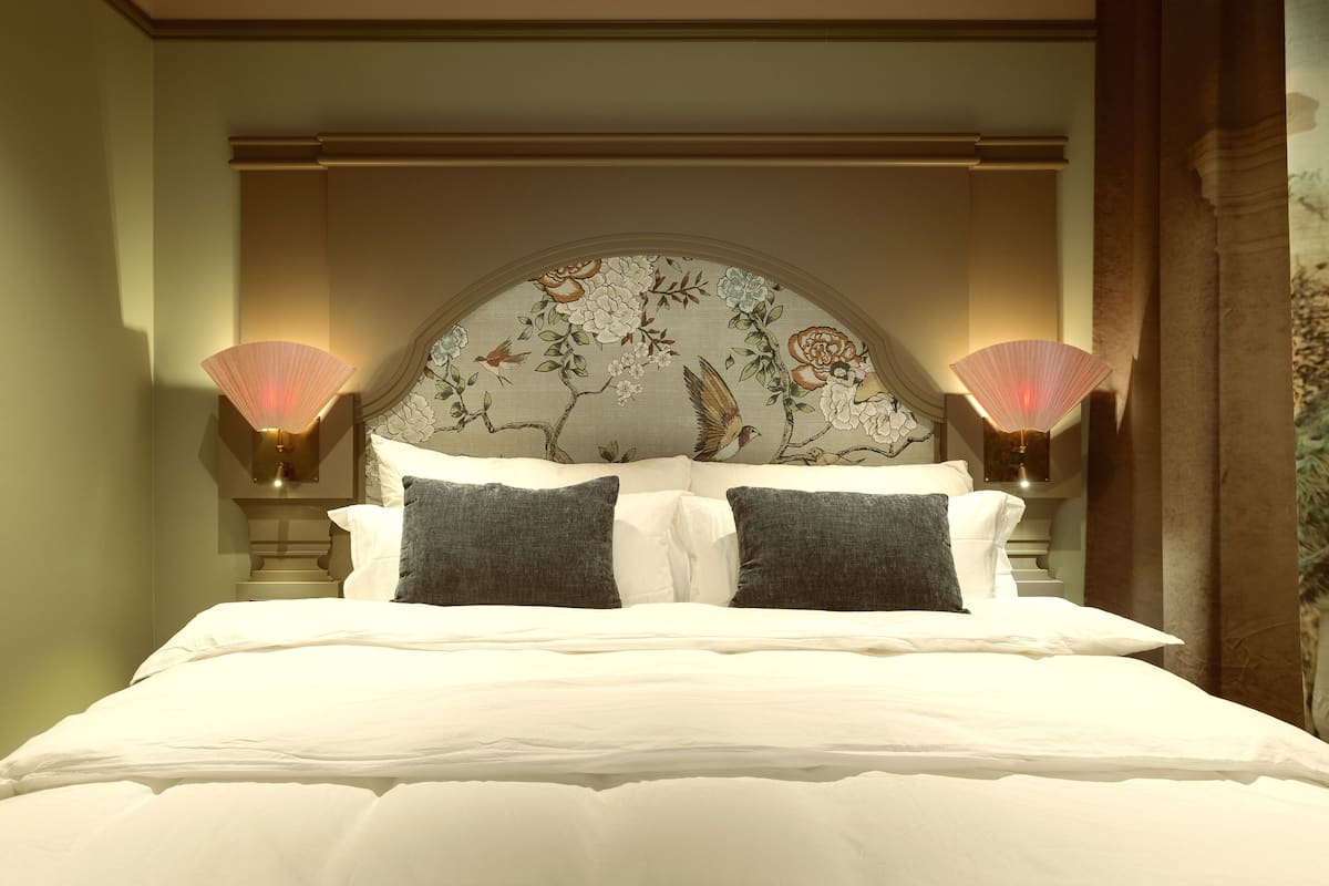Un lit bien fait, avec du linge de lit blanc et deux oreillers foncés, flanqué de deux lampes murales à motifs floraux.