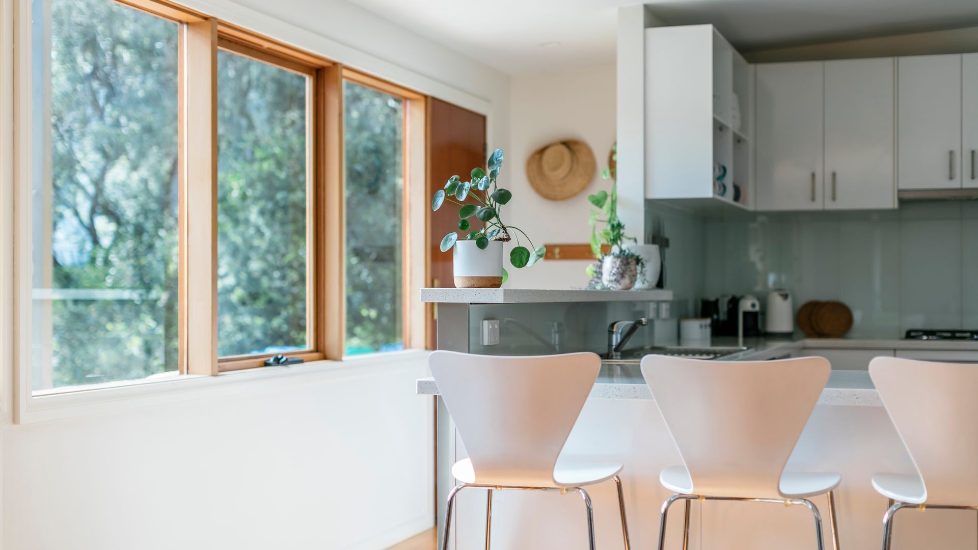 Una cocina moderna con armarios blancos, un comedor con sillas blancas y un amplio ventanal con vistas a árboles.