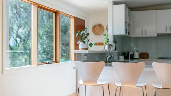 A modern kitchen with white cabinets, a dining area with white chairs, and a large window with a view of trees.