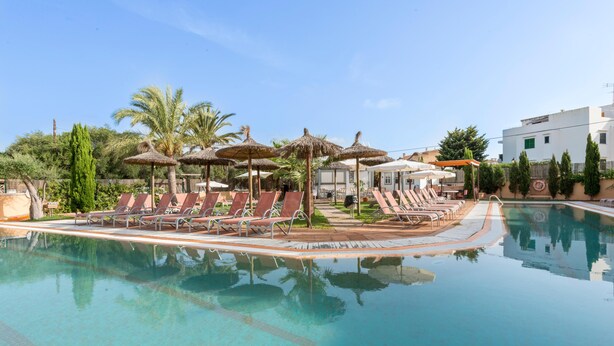 A swimming pool with lounge chairs and umbrellas, surrounded by palm trees and a building. - Playa Ferrera Apartments (Felanitx)