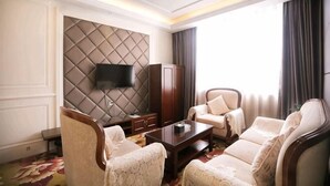 Quarto de hotel com uma televisão de tela grande fixada em parede acolchoada, um conjunto de sofá, uma mesa de centro de madeira e uma janela grande com cortinas.